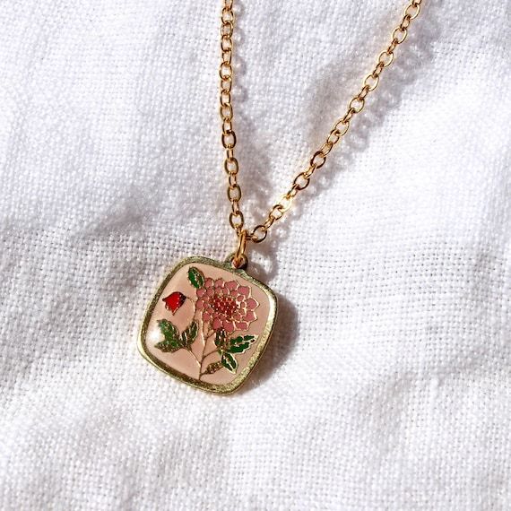 Ett vintage guldhalsband med ett kvadratiskt, blommigt emaljhänge på vitt tyg.